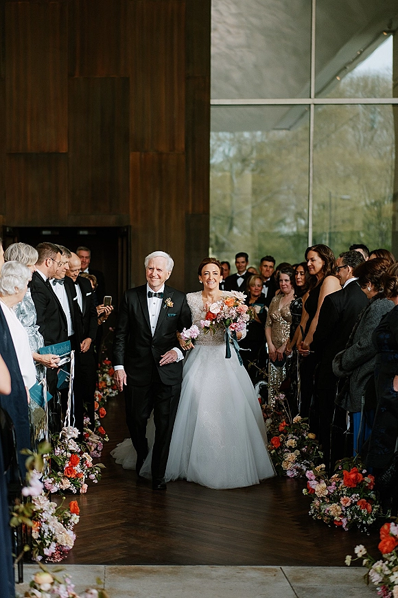 Wedding processional as bride walking down aisle with her father, holding a coral and pink bouquet in a wood-and-glass indoor venue