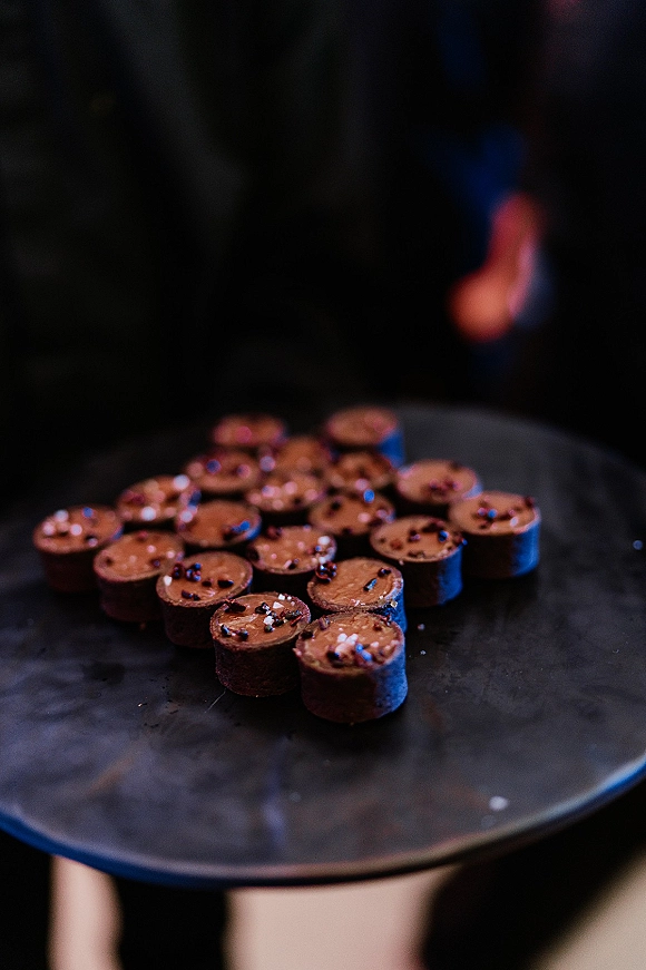 Wedding dessert bites on a round serving tray, mini chocolate desserts topped with sprinkles, against a dark backdrop with blurred guests