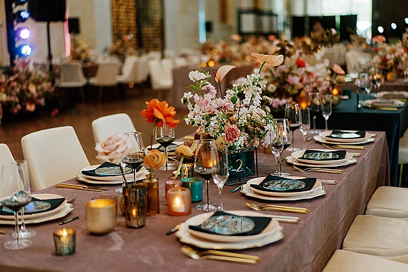Reception tablescape wedding table decor with floral centerpieces, candles, gold flatware, and upholstered chairs in an ambient-lit hall