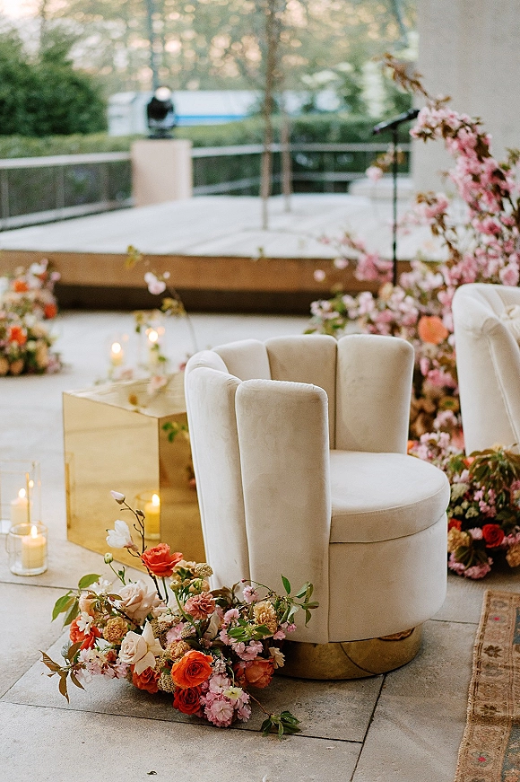 Ceremony lounge seating with cream velvet chairs, gold pedestal side table, floral clusters and glass cylinder candles on an outdoor patio stage