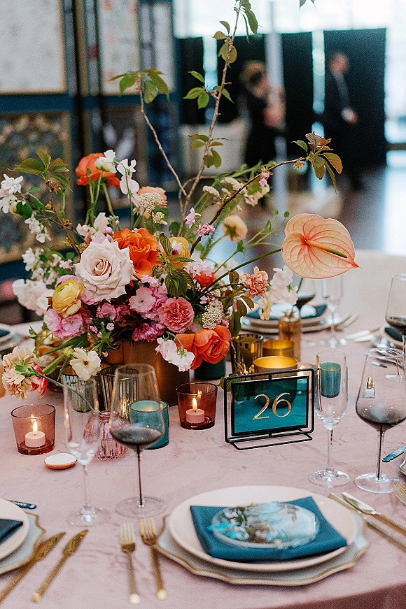 Reception tablescape with a wedding table centerpiece of anthurium, roses, and greenery branches, votive candles, and glassware in an indoor venue