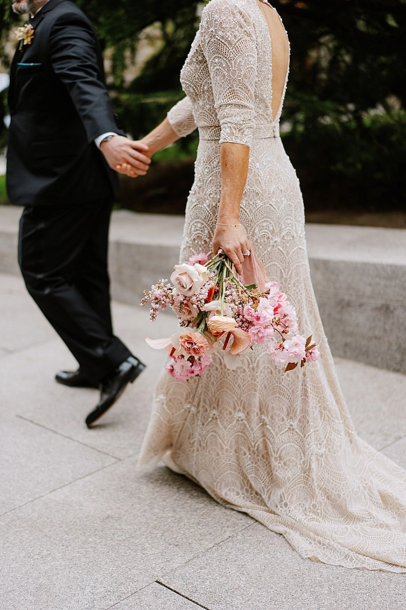 Couple portrait of bride and groom holding hands, bride in lace wedding dress with pink bouquet on stone walkway by trees