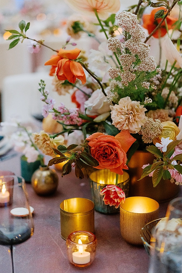 Reception centerpiece with wedding floral centerpiece of orange roses, blush blooms, greenery, and candles in gold and amber votives on a bokeh-lit table