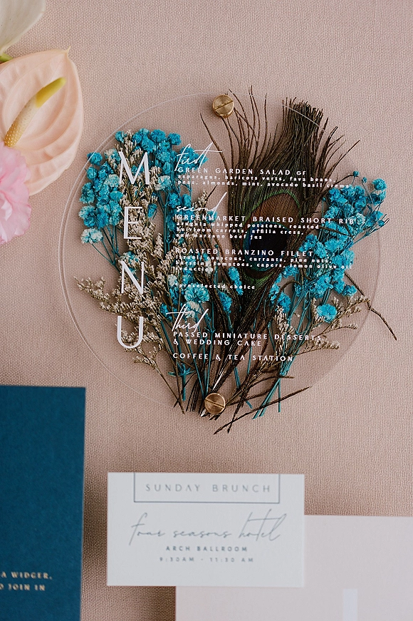 Wedding menu card on clear acrylic with dried blue flowers and a peacock feather, secured by brass fasteners on neutral linen fabric