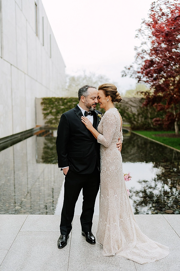 Couple portrait of bride and groom embrace with a forehead touch, her lace dress and bouquet by a reflecting pool near modern building