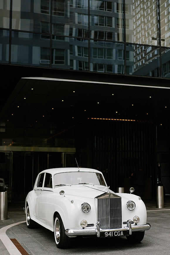 Wedding getaway car, a vintage white car with chrome grille and round headlights parked under a modern hotel canopy on a city street