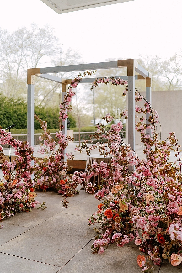 Wedding ceremony arch with floral ceremony arch of pink blossoms, roses, anthurium, and greenery on a wooden frame on stone patio with mic stand