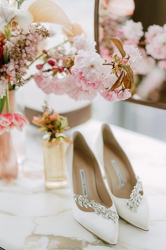Bridal shoes, white wedding heels with crystal embellishments beside pink flowers and perfume on a marble tabletop by a mirror