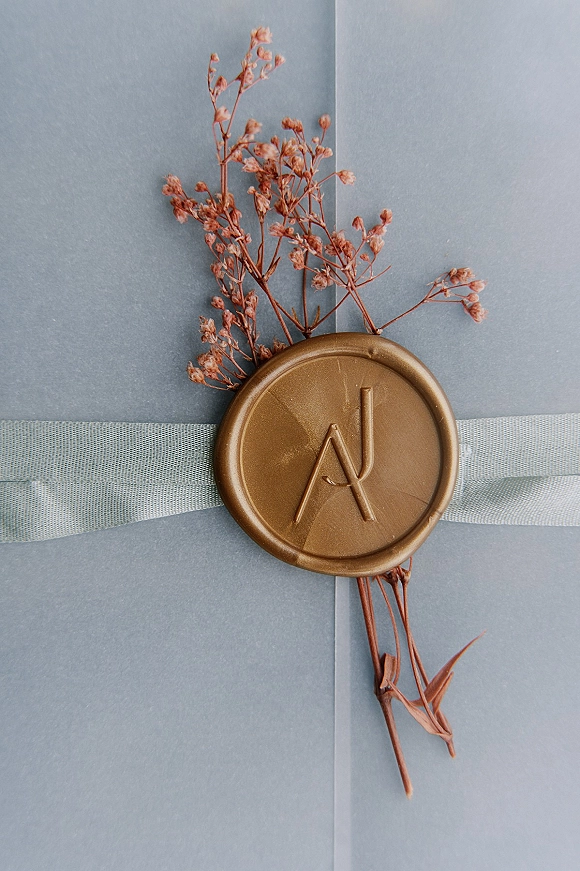 Wax seal invitation with wedding invitation wax seal, monogram stamp, ribbon, and dried flowers on a blue-gray envelope flat lay