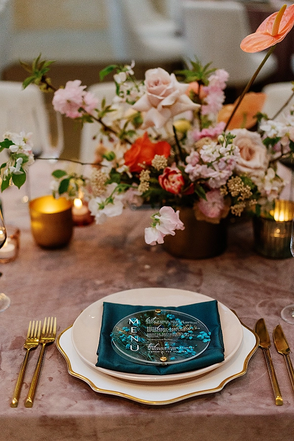Reception tablescape with wedding table centerpiece of roses, sweet peas and anthurium, gold flatware, teal napkin and votive candles in a softly lit room