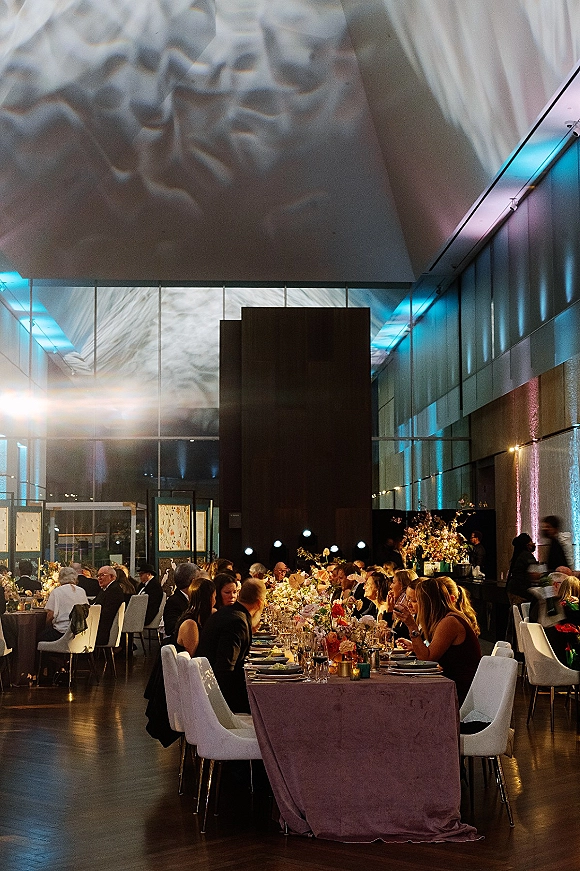 Wedding reception decor on long banquet tables with velvet tablecloths, floral centerpieces and taper candles in a modern high-ceiling hall