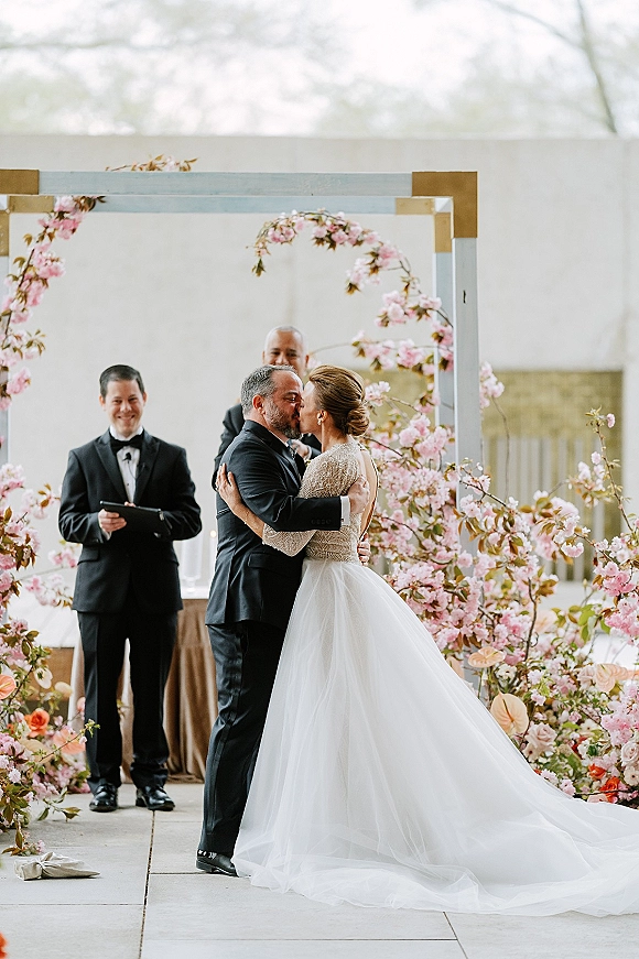 Wedding kiss under a pink blossom floral arch, bride in lace gown and veil with groom in tux on an outdoor terrace altar