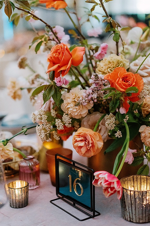 Reception tablescape with wedding table centerpiece of orange roses, pink blooms and greenery, plus votive candles and gold table number in bokeh-lit room