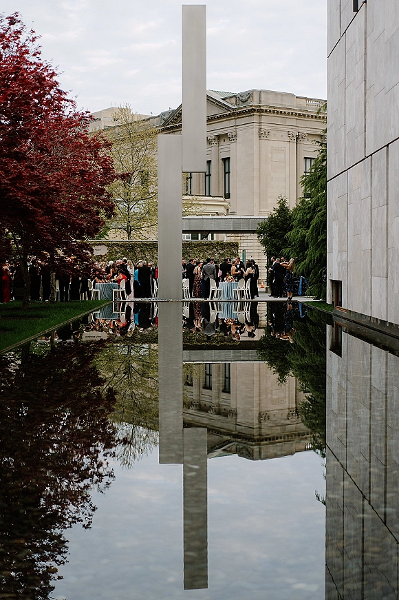 Wedding cocktail hour with guests mingling around high-top tables with linens and floral centerpieces beside a reflecting pool in a museum courtyard