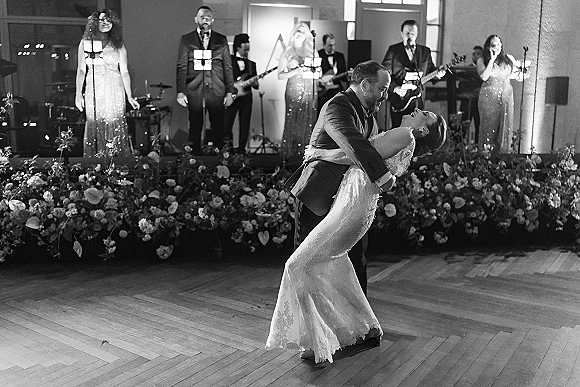 First dance as groom dips bride in lace wedding dress on reception dance floor, live band on stage under bright lights and florals