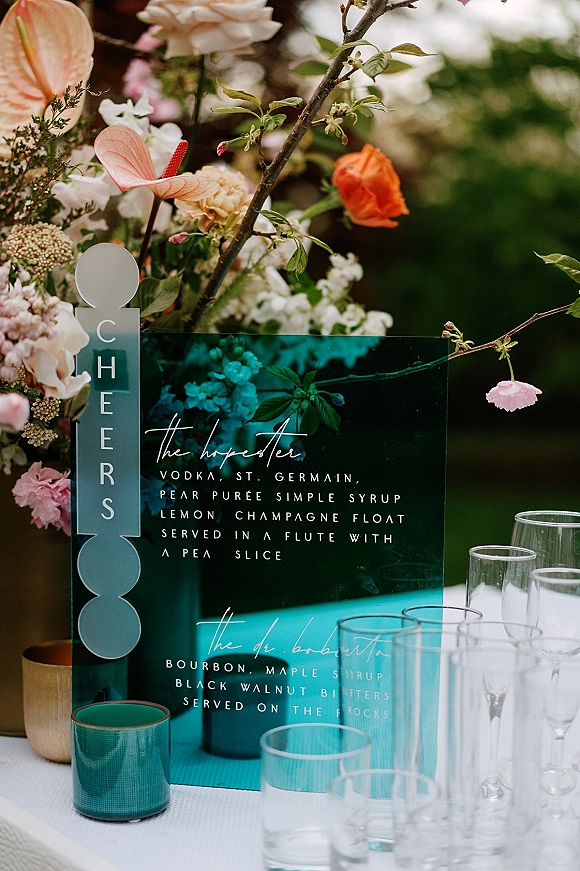Wedding bar signage with acrylic cocktail menu text beside anthurium and roses, teal glass votive and flutes on a garden bar table