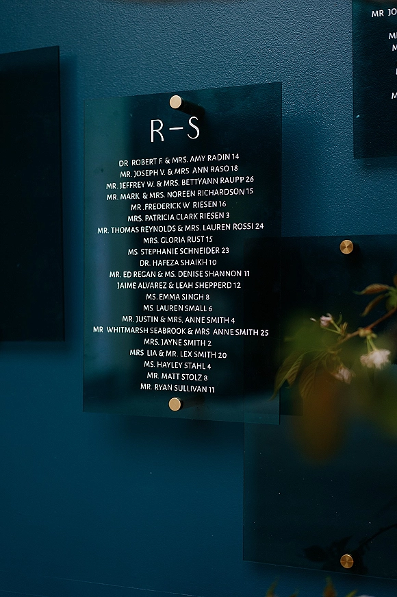 Wedding seating chart on a clear acrylic sign with white lettering and gold standoffs, wall-mounted against a dark blue wall with greenery