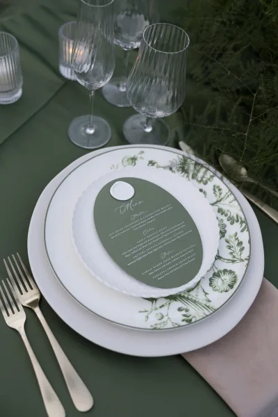 Reception place setting with wedding tablescape details, layered ceramic plates and a sage green menu card on a green tablecloth with ribbed glasses