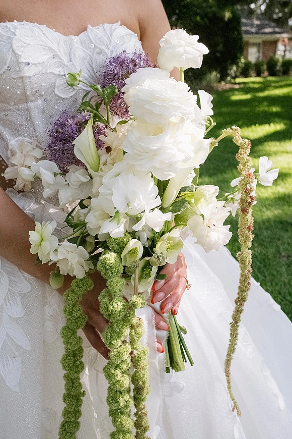 Bridal bouquet of white and purple blooms with cascading floral strands, held by bride in strapless lace gown on sunlit lawn with trees