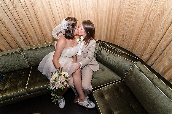 Wedding kiss portrait of a lesbian couple on a green velvet couch, one in a suit and sneakers, the other in a veil holding a bouquet, with a gold curtain backdrop