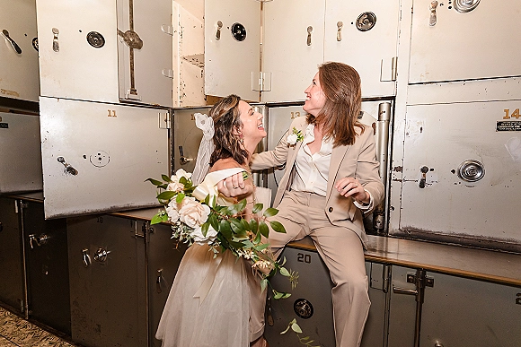 Couple portrait of a same sex wedding couple leaning together, one holding white rose bouquet, in front of industrial lockers indoors