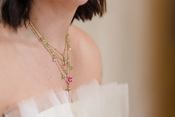 Bridal necklace with layered necklace bride styling, gold chains and green bead charms over a strapless tulle wedding dress on a warm wall
