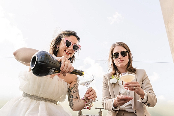 Wedding toast moment as the bride pouring champagne into coupe glasses beside the groom, both in sunglasses on an outdoor patio railing against sky