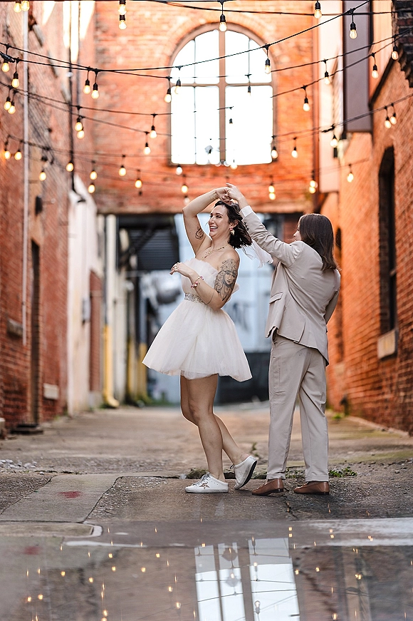 Couple dancing under string lights as the bride twirls in a short wedding dress and sneakers beside a brick alley puddle reflection