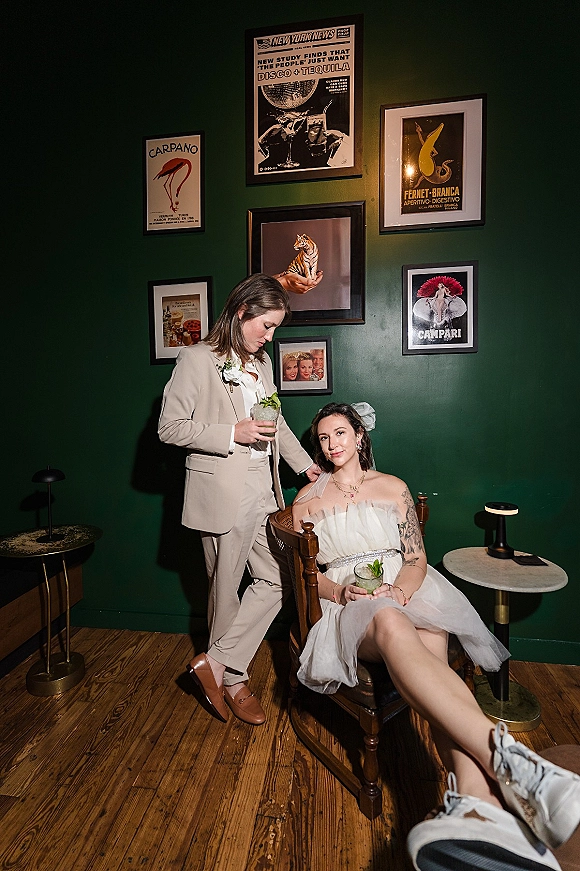 Couple portrait of two brides in short dress and suit holding cocktails, one seated in a rocking chair before a dark green gallery wall
