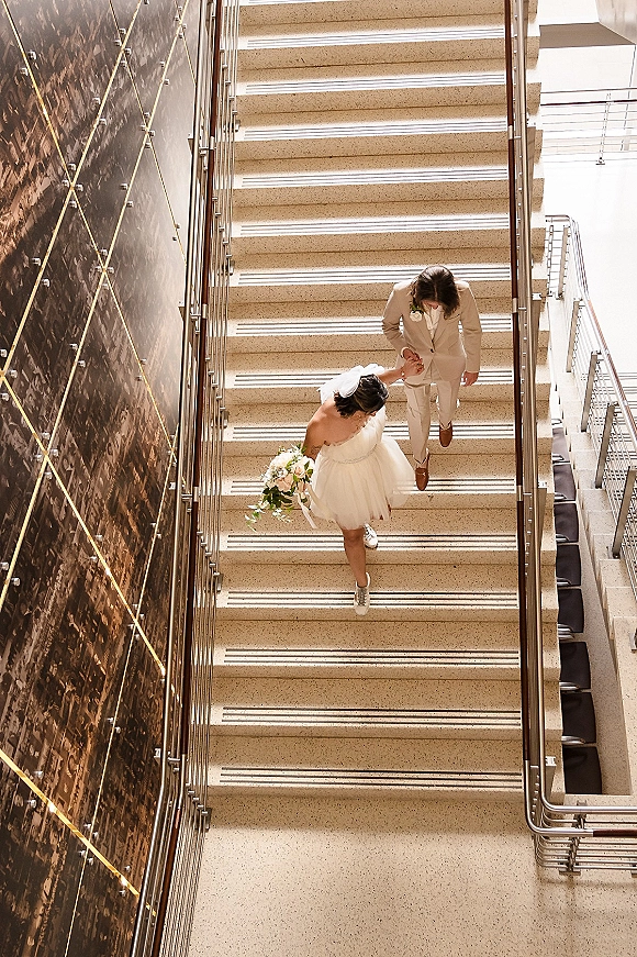 Couple portrait of bride and groom on stairs holding hands, her short wedding dress and bouquet visible in top-down view indoors