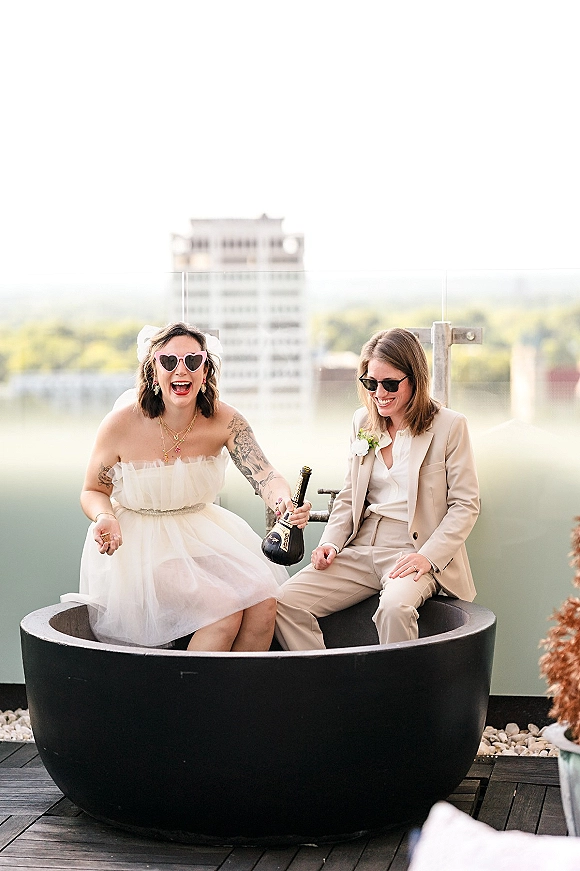 Couple portrait of a lesbian wedding couple in a bathtub, one in a short dress and one in a beige suit, with city skyline behind
