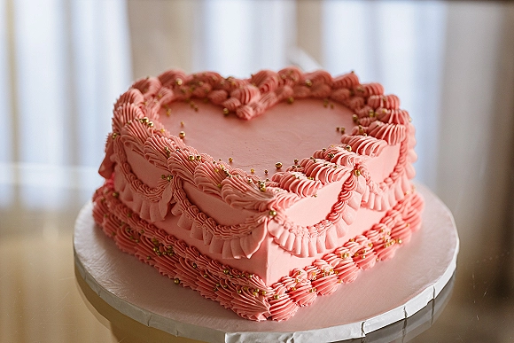 Wedding cake in a heart shaped wedding cake design with pink ruffle buttercream, gold dragees, on a tabletop in window light near curtains