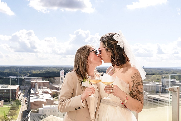 Wedding kiss between a lesbian couple holding champagne coupe glasses on a rooftop terrace with city skyline, blue sky, and veil accents