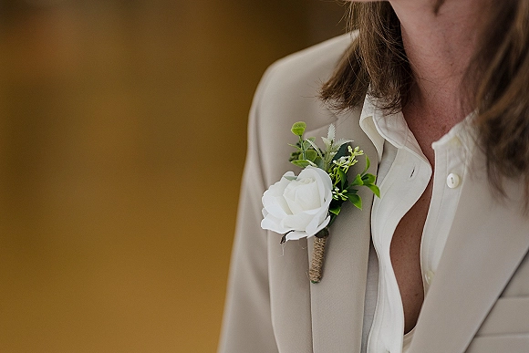 Wedding boutonniere with a white rose boutonniere and greenery, twine-wrapped on a beige suit lapel against a plain wall