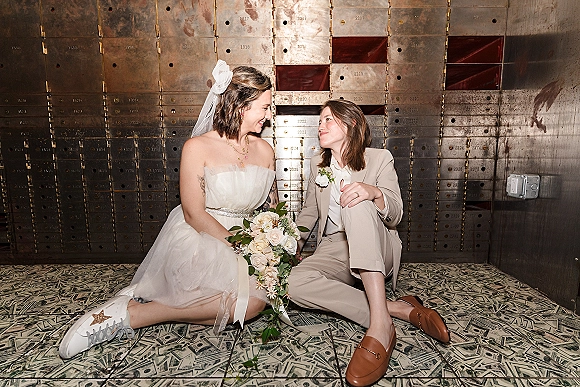 Couple portrait of brides sitting on a money-print floor, one in a wedding dress and sneakers holding a bouquet, posed by a bank vault door