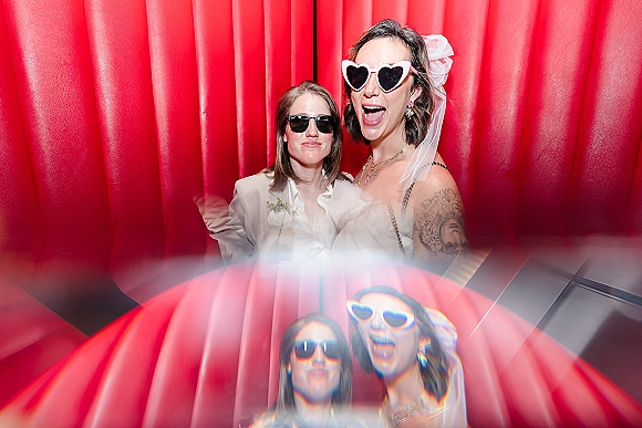 Wedding photo booth moment with two women in heart sunglasses, one in a veil, posing by a mirror against a bold red curtain backdrop