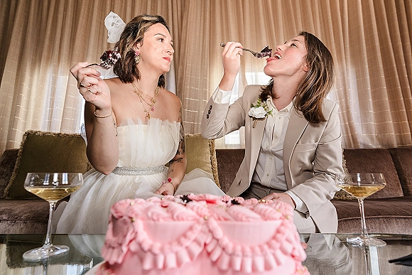Wedding cake moment as two brides share bites of a pink ruffle cake on a couch, champagne coupes on a glass table in window light