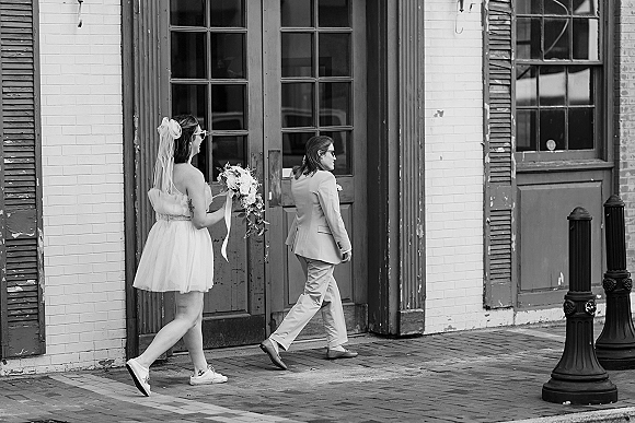 Wedding couple portrait in black and white, walking on a city sidewalk past a brick facade; bride in short dress with veil bow, ribbons, sneakers, both in sunglasses