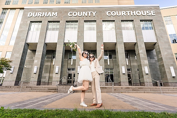 Couple portrait at a courthouse wedding photo, raising bouquets with arms up; one in short dress and sneakers, other in beige suit and sunglasses