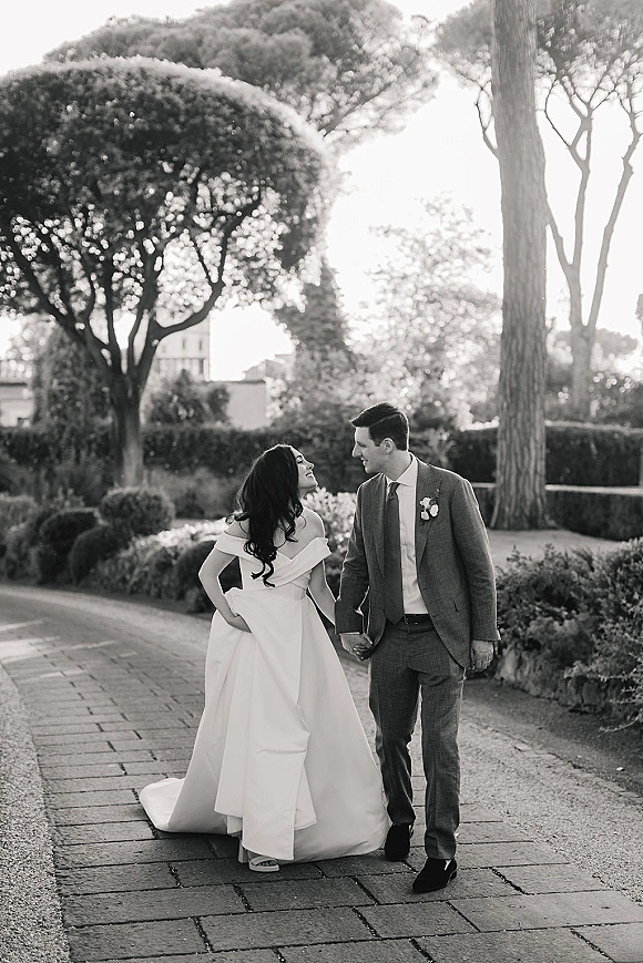 Couple portrait of bride and groom walking hand in hand on a garden path, her off-the-shoulder gown trailing as he wears a suit boutonniere