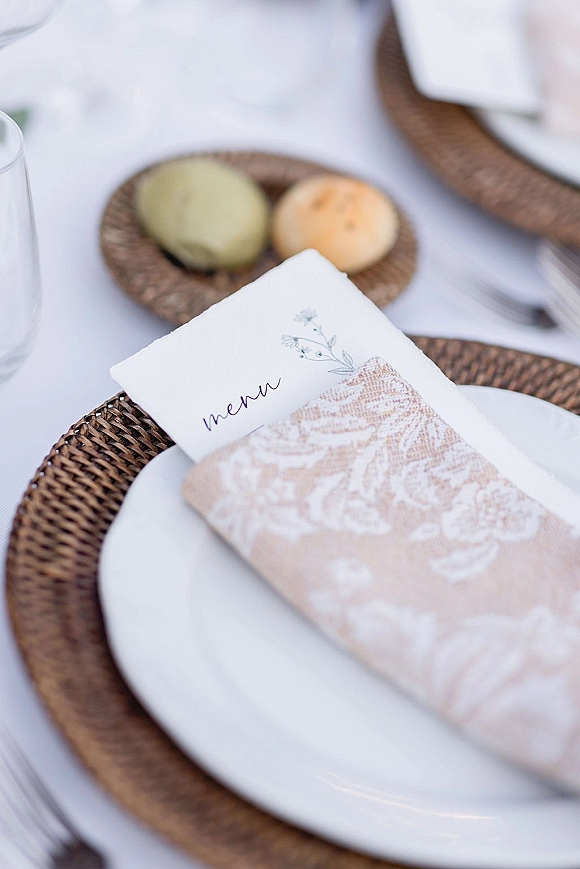 Reception place setting with wedding menu card on a white plate atop a rattan charger, linen napkin, flatware, glass, and bread rolls on a white tablecloth