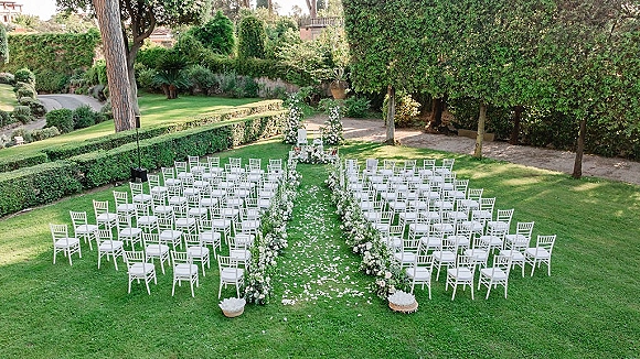 Ceremony setup for an outdoor wedding ceremony with white chairs lining a floral aisle, rose petals on a runner, on a garden lawn path