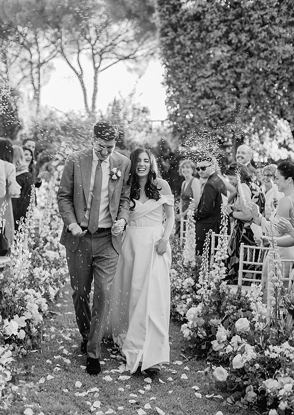 Wedding recessional as bride and groom walking down aisle holding hands, guests cheering beside petal-strewn path in garden trees