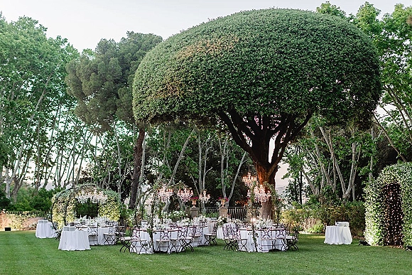 Outdoor reception decor with garden wedding reception tables under a tree canopy, crystal chandeliers, candles, florals, and string lights by a hedge wall