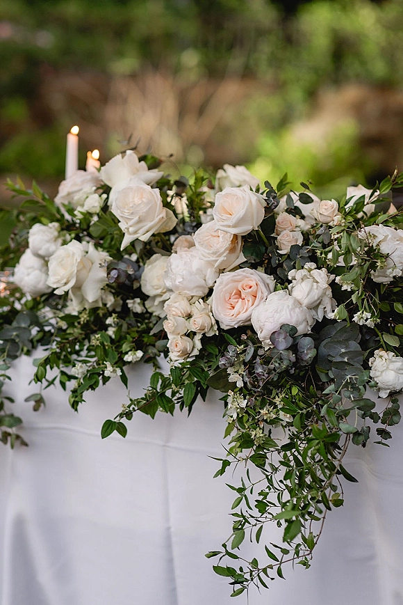 Wedding floral centerpiece with blush and white roses and eucalyptus, arranged beside taper candles on a white tablecloth in a blurred garden setting