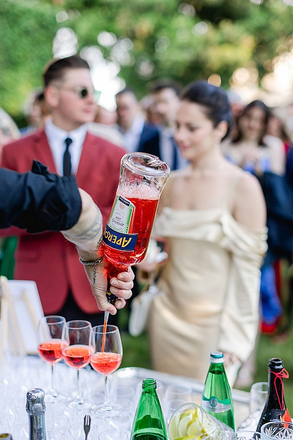 Wedding cocktail hour with wedding aperol spritz setup, wine glasses, aperitif bottle, lemons, and ice bucket on a garden table among guests