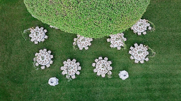Outdoor reception setup with round wedding tables in white tablecloths, floral centerpieces and greenery garlands on a grass lawn by a hedge