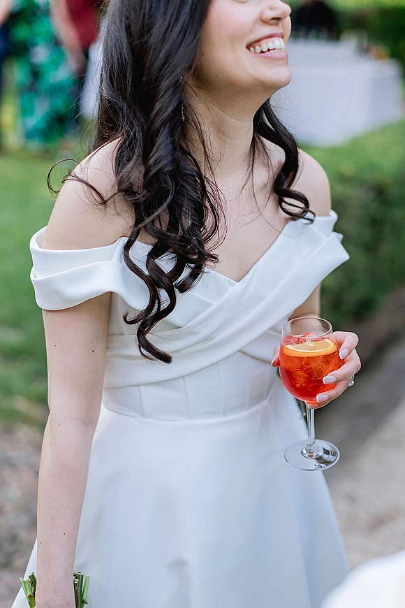Bride portrait in an off shoulder wedding dress, laughing with a cocktail glass and orange slice garnish at a garden reception with blurred guests