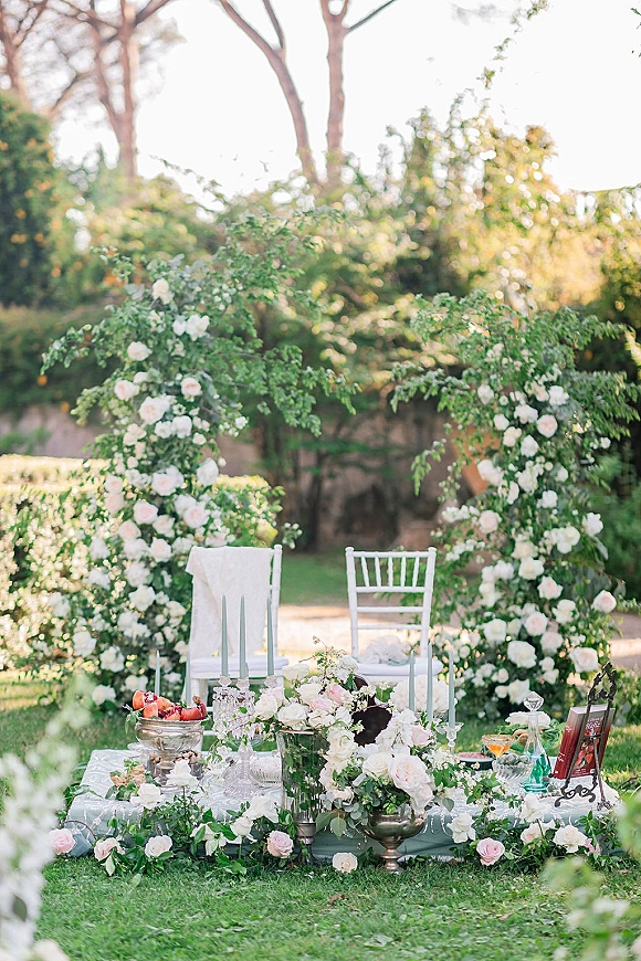 Outdoor reception tablescape with a low picnic wedding table, rose and greenery garlands, taper candles and crystal glassware on a garden lawn