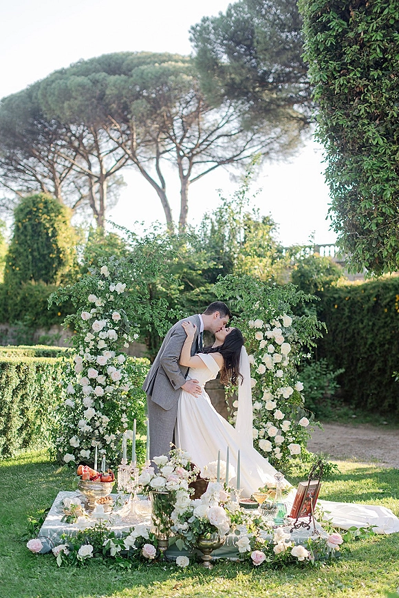 Wedding kiss portrait of bride and groom kissing beneath a white rose and greenery floral arch, veil flowing in a lush garden setting